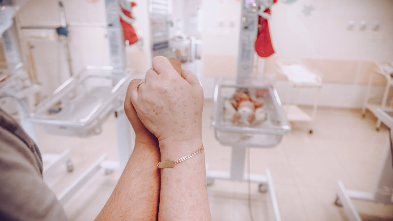 A parents hands clasped together in a hospital nursery, with newborns in the background.